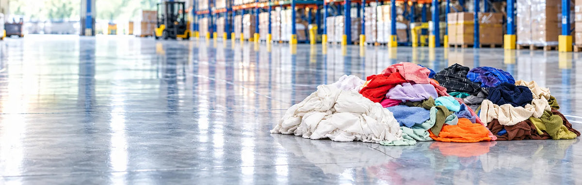 A pile of assorted colorful clothing items sits on a polished warehouse floor, with shelving and boxes visible in the blurred background.